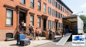 People carrying moving boxes down the steps of a red-brick rowhouse and loading them into a moving truck parked on a narrow city street, with moving supplies nearby.